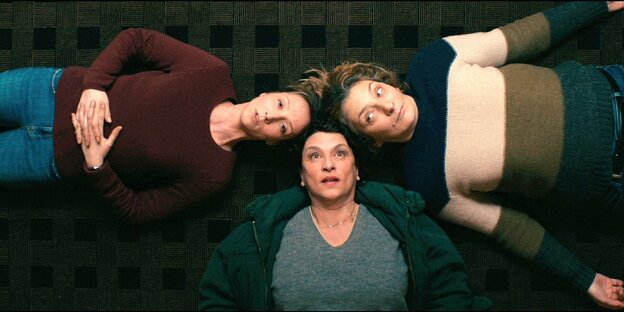 Fotoblick von oben, drei Frauen liegen Kopf an Kopf auf dem Teppich.