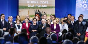 Menschen stehen neben- und hintereinander auf einer Bühne. Auf einem Plakat im Hintergrund steht: „Ein guter Moment für Polen“