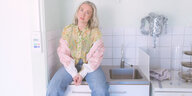 Eine blonde junge Frau in Jeans und einer bunten Bluse sitzt auf einer Küchenspüle