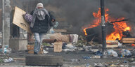 ein vermummter Mann mit einem selbstgebastelten Schild auf einer vermüllten Straße, im Hintergrund brennt es