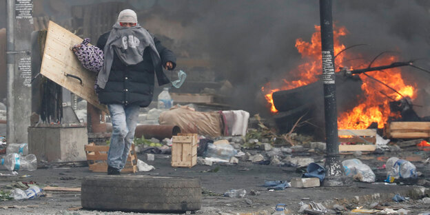 ein vermummter Mann mit einem selbstgebastelten Schild auf einer vermüllten Straße, im Hintergrund brennt es