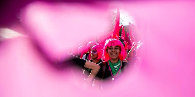 Durch ein Loch in einem rosafarbenen Transparent sieht man eine junge Frau mit einen grünen Halstuch auf einer Demonstration.