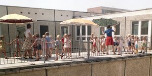 Kinder und Erzieherinnen unter Sonneschirmen auf der Terrasse eines Plattenbaus