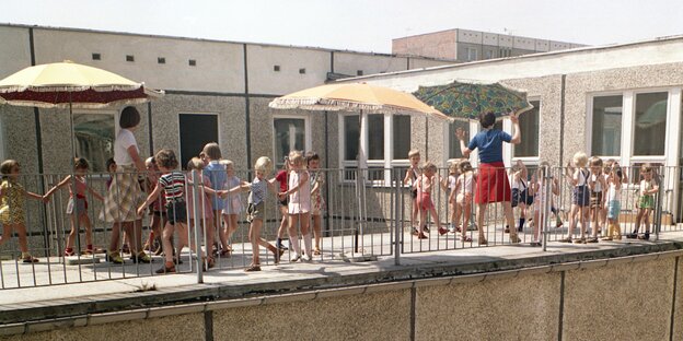 Kinder und Erzieherinnen unter Sonneschirmen auf der Terrasse eines Plattenbaus Kinder und Erzieherinnen unter Sonneschirmen auf der Terrasse eines Plattenbaus