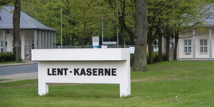 Schriftzug Lent-Kaserne auf weißem Schild, Wiese, dahiner weiße Kasernengebäude.