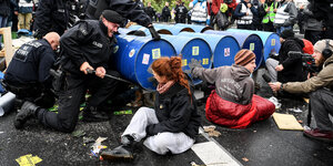 Polizisten entfernen eine aus Metallfässern errichtete Blockade, während AktivistInnen daneben sitzen