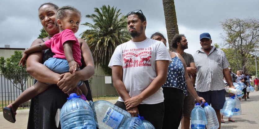 Menschen stehen in einer Reihe. Sie tragen mindestens zwei leere Wasserkanister in der Hand. Die Kanister haben ein Fassungsvermögen von fünf Litern