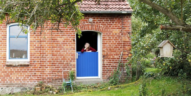 Ein Backsteinhaus im Grünen, eine Frau schaut aus dem Fenster