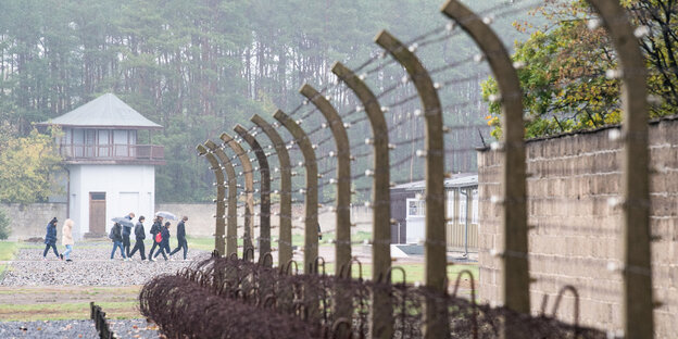 Besucher gehen über das Gelände der Gedenkstätte Sachsenhausen, im Vordergrund ist ein Zaun aus Stacheldraht zu sehen