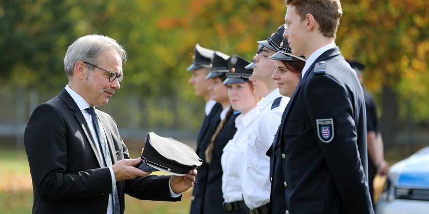 ein Mann im Anzug steht vor einer Reihe Polizisten und hält die Mütze eines der Polizisten in den Händen