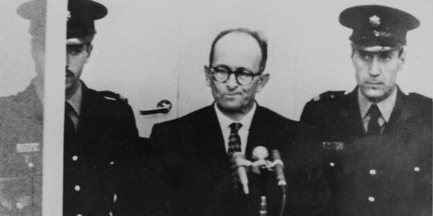 Zwischen zwei Uniformierten steht Eichmann im Anzug mit Krawatte vor einem Mikrofon.