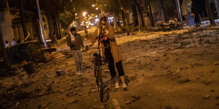 Eine Frau hält sich ein Tuch vor die Nase und schiebt ihr Fahrrad über eine Straße voller Schutt