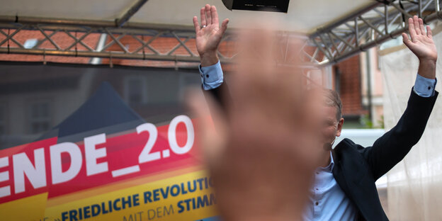Eine Hand im Vordergrund verdeckt das Gesicht eines winkenden Mannes auf einer Wahlkampfbühne.