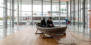 In einem Pavillon, hinter dessen Fenstern man Schiffe sieht, steht ein Boot auf dem Parkett. Zwei Männer mit obskuren Brillen sitzen darin.