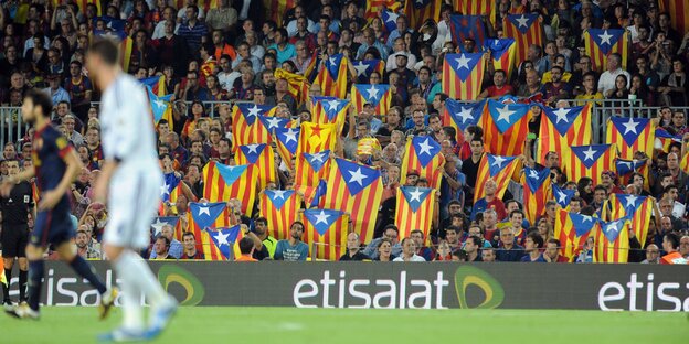 Man sieht im Vordergrund verschwommen zwei Fußballspieler auf der Stadionwiese, im Hintergrund viele Barcelona-Fans, die katalanische Flaggen hochhalten.