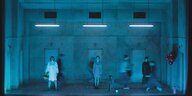 Eine Bühne in kaltes blaues Licht getaucht, drei Frauen stehen dort, andere Figuren verlieren sich in der Bewegungsunschärfe.