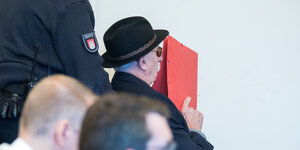 Ein Mann wird mit einem roten Aktendeckel vor dem Gesicht in den Gerichtssaal gebracht