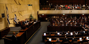 Die Knesse. Vor einer Steinwand sitzen auf einem Podium mehrere Menschen. Hinter ihnen sind Flaggen Israels. Vor ihnen sitzen sie Abgeordneten in mehreren Reihen in U-Form