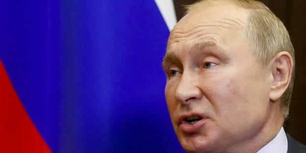 Vladimir Putin, ein alter Mann mit wenigen Haaren. Hinter ihm eine Russlandfahne