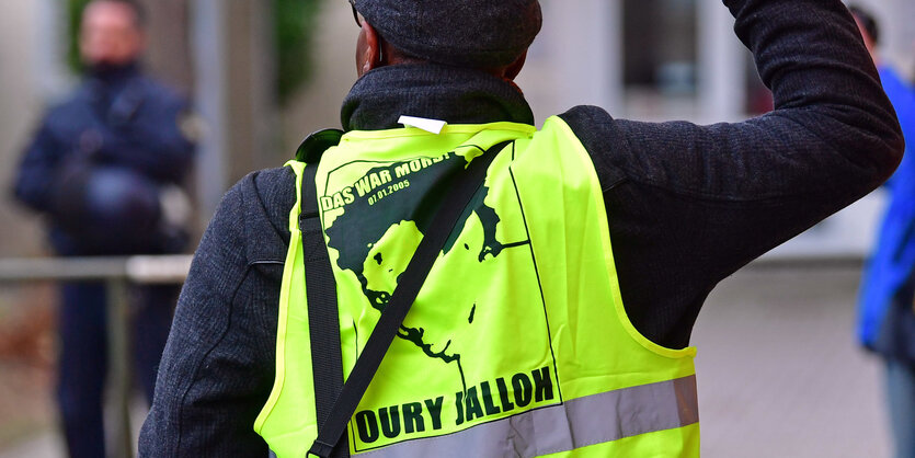 Ein Teilnehmer einer Demonstration in Gedenken an Oury Jalloh wendet sich in Dessau-Roßlau an Polizisten. Er trägt eine Warnweste mit dem Gesicht Jallohs darauf und dem Satz" Das war Mord"