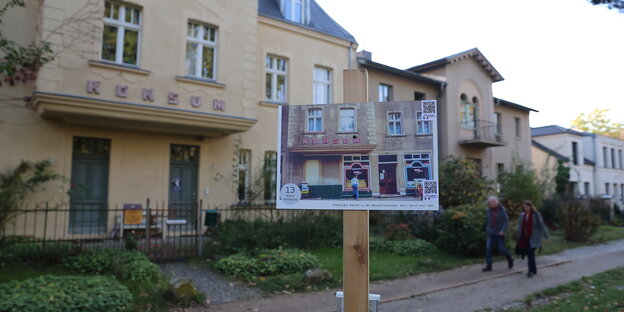 ein Wohnhaus auf dem „Konsum“ steht, davor eine Fototafel, die den Konsum zur Zeit des Betriebes zeigt