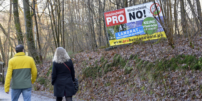 Spaziergänger gehen an einer Plakatwand mit der Aufschrift "Holzlandwald Pro Windradwald No! Landrat, wir nehmen sie beim Wort! Keine Windräder im Wald!" im Mühltal im Thüringer Holzland vorbei