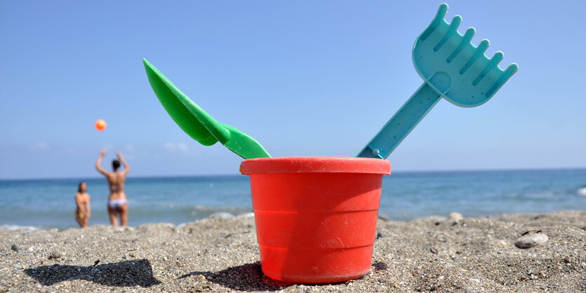 Ein roter Plastikeimer mit Schaufel und Hacke darin steht am Strand