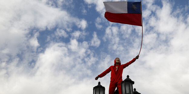 Ein Demonstrant in Santiago de Chile trägt einen roten Ganzkörperanzug und eine Guy-Fawkes-Maske, steht auf einer Straßenlaterne und hält die chilenische Flagge in die Höhe