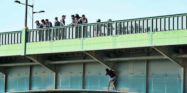Eine Brücke. Auf ihr stehen Polizisten, die die Straße sperren. Auf dem Brückengerüst, direkt unter den Polizisten klettert ein Mann entlang, um die Brücke dennoch überqueren zu können