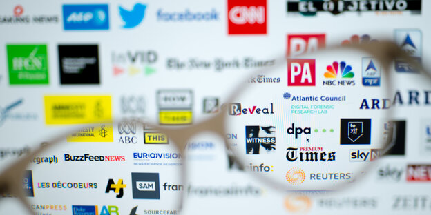 Eine Brille wird vor diverse Medien-Logo gehalten, darunter Twitter, Facebook, CNN und dpa