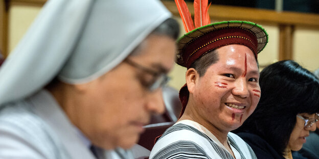 Im Vordergrund eine Nonne, im Hintergrund ein lächelnder Indigener aus Brasilien mit Gescihtsbemalung und Federschmuck: Auf der Amazonas-synode im Vatikan