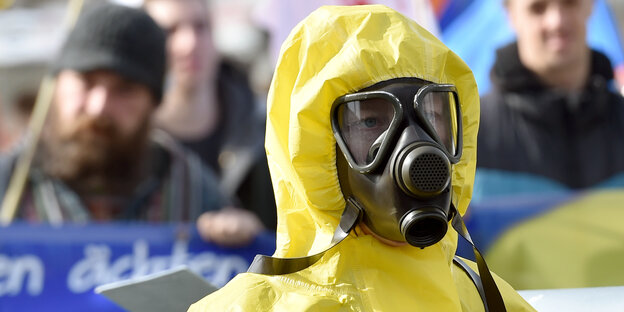 Eine Frau demonstriert mit einer Atemschutzmaske und gelbem Schutzanzug