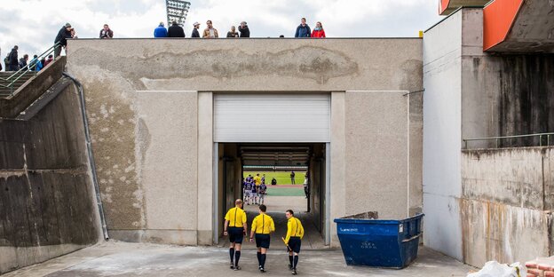 Schiedsrichter laufen in ein Stadion an einem großen Bauschuttcontainer vorbei