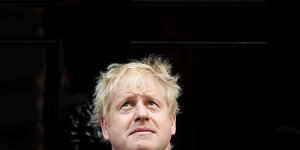 Boris Johnson steht vor einer schwarzen Tür und guckt nach oben