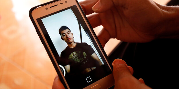 Hände halten ein Handy, auf dessen Bildschirm das Foto eines jungen Mannes zu sehen ist.