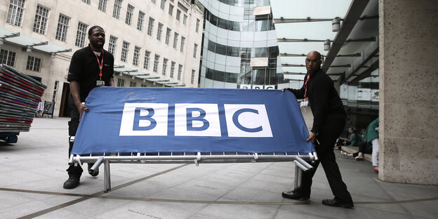 Zwei Männer tragen eine Absperrung, die mit BBC beschriftet ist.