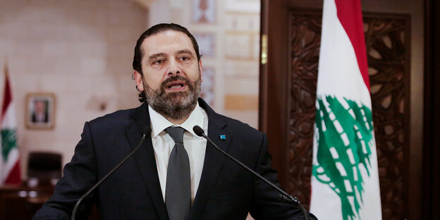 Ein Mann in Anzug steht vor Mikrophonen und hinter der libanesischen Flagge.