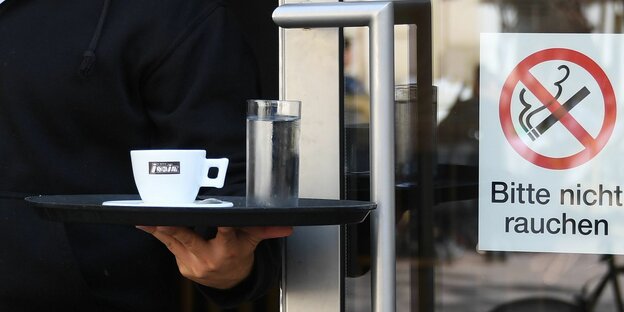 Kellner trägt ein Tablett mit Kaffee und Wasser. An der Tür steht: Bitte nicht rauchen!