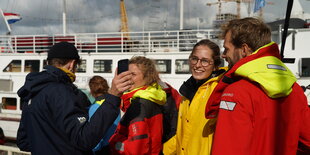 Vier Menschen auf einem Schiff. Die Person links hält ein Handy in die Höhe, die mittigen Personen lachen in die Kamera. Von der Person rechts im Bild sieht man nur den Rücken.