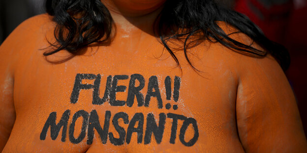 „Raus!! Monsanto“, steht auf der Brust einer Frau mit nacktem, orange angemaltem Oberkörper