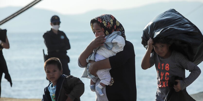 Eine Frau mit Baby in Arm und zwei Jungen um sich herum vor einem Hintergrund mit Wachleuten und Meer.