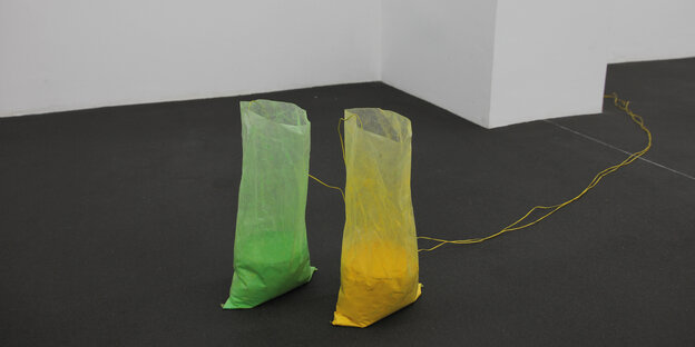 Zwei Plastiktüten mit grünem und gelbem Pigment stehen im auf dem Fußboden im Raum, zwei je gelb und grün besprühte Lautsprecher stecken in ihnen, dazu kommen Kabel und ein CD-Spieler