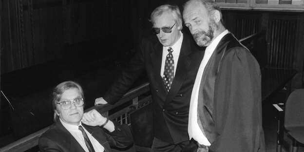 Drei Männer stehen und sitzen in einem Gerichtssaal