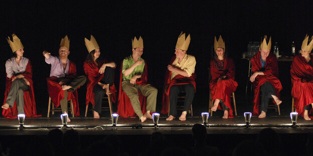 Acht Frauen und Männer sitzen auf Stühlen nebeneinander, in rote Umhänge gehüllt und mit Pappkronen, auf der Bühne.