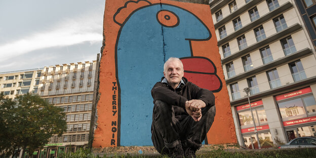 Der Künstler Thierry Noirm ein älterer, weißhaariger Mann, sitzt vor einem bemalten Mauerstück.
