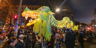 Abends auf einem Platz, viele Menschen tragen einen grün leuchtenden Drachen, Häuser im Hintergrund.