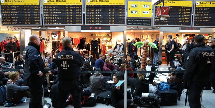 Demnonstranten sitzen hinter Absperrbändern auf dem Boden, beobachtet von Polizisten