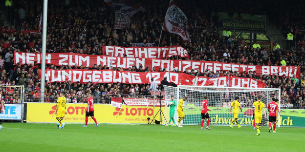 Freiburg Fans mit enem großen Transparent, auf dem Rasen eine Torszene