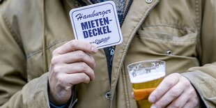 Ein Mensch in beiger Jacke hält in der einen Hand ein Getränk, in der anderen einen Bierdeckel mit der Aufschrift Hamburger Mietendeckel