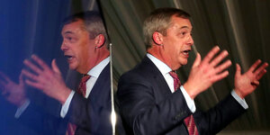 Nigel Farage gestikuliert und wird in einer Scheibe gespiegelt